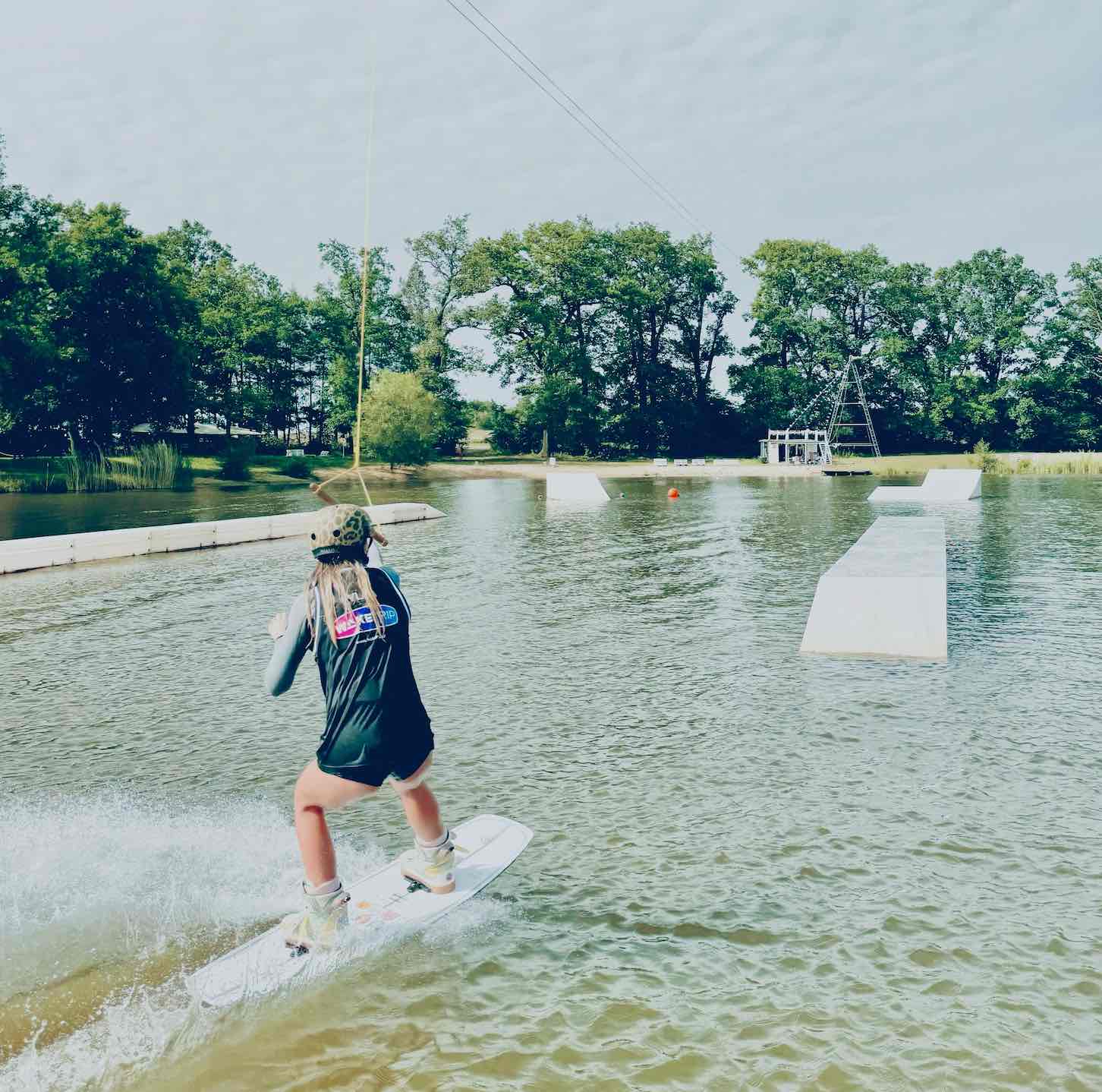 Wakeboarder in a Wake Trip-branded shirt riding behind the cable tow past kicker ramps, with the start dock and tower visible across the lake